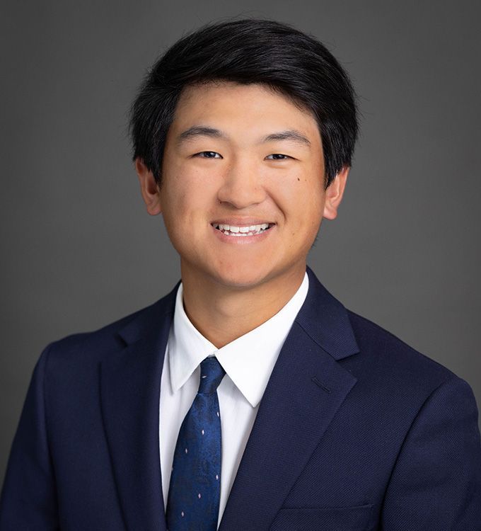 Studio headshot of a smiling person in a navy suit and tie against a gray background