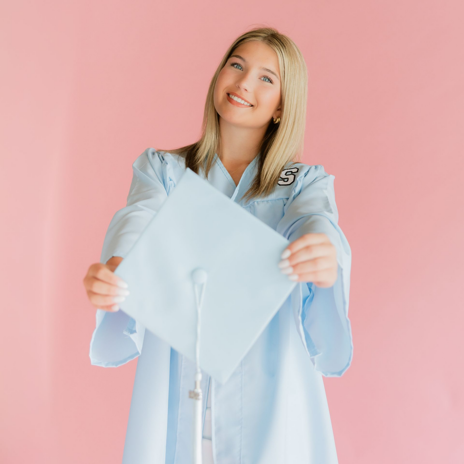 Smiling graduate in a light blue gown holding a white cap against a pink background