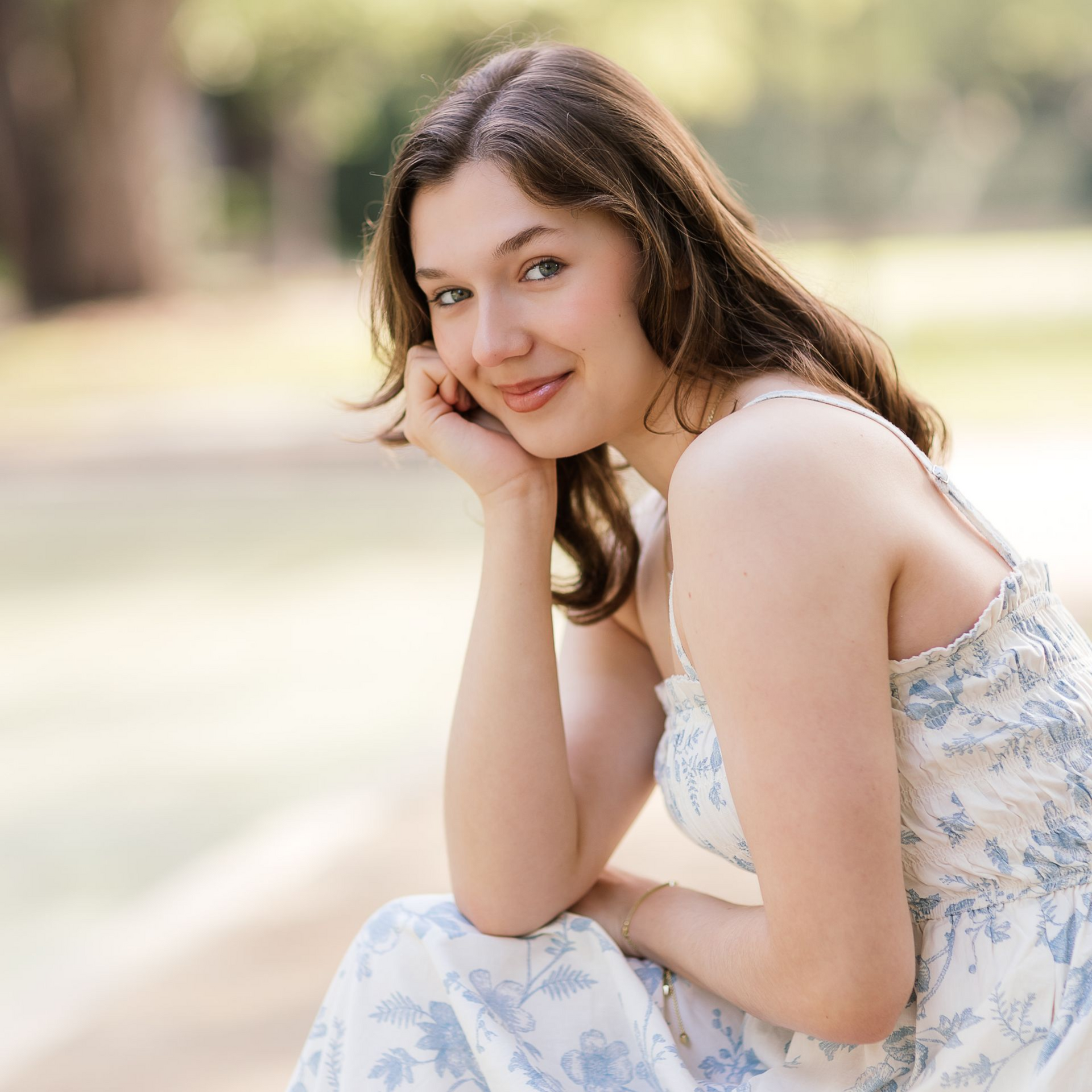 Young woman in a floral dress sitting outdoors, smiling softly with her head resting on her hand
