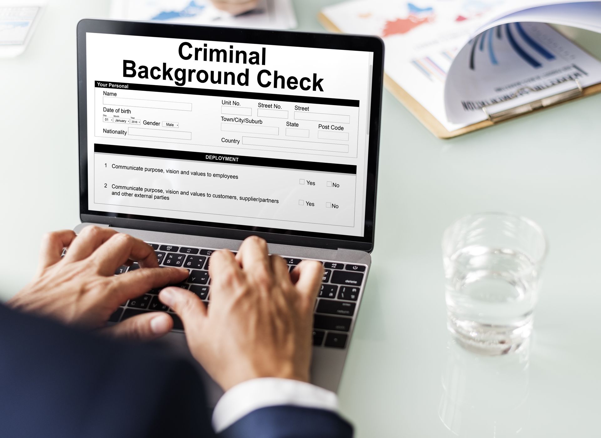 Hands typing on laptop displaying a criminal background check form. A glass of water sits nearby.