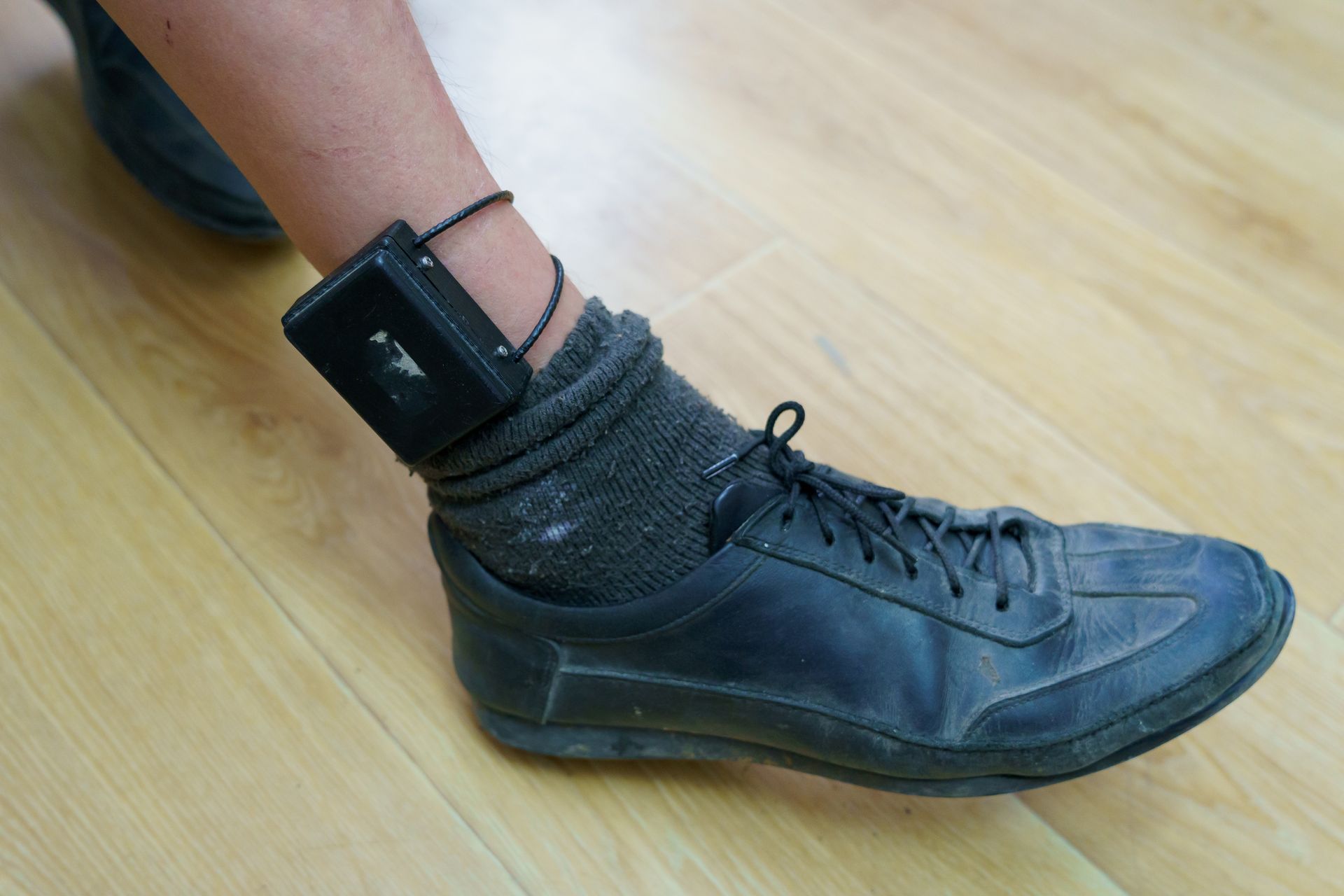 Ankle of person wearing a black ankle monitor secured above a black shoe.