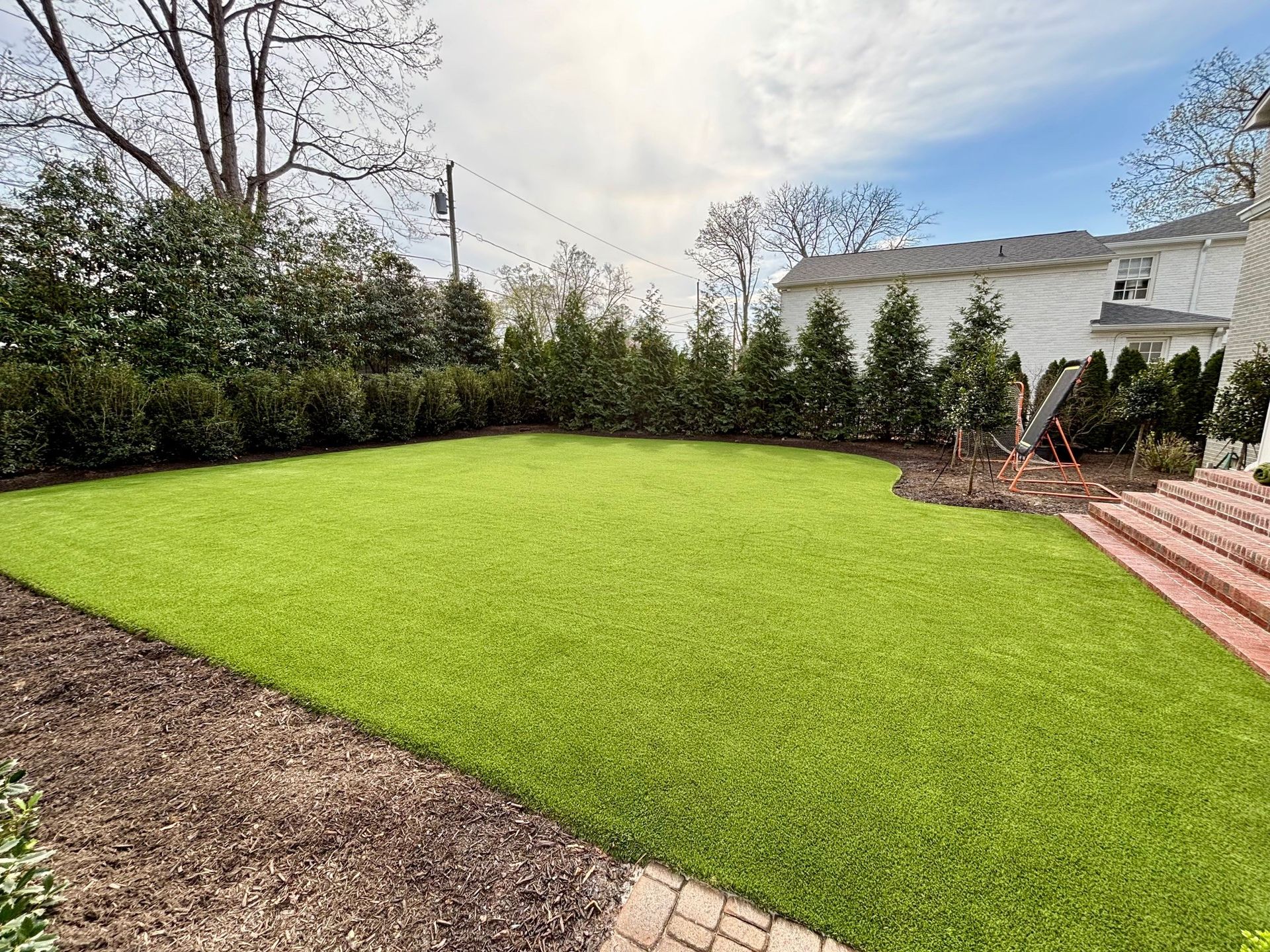 A neatly manicured artificial lawn in a backyard, surrounded by a hedge, with brick stairs on the right and mulch borders.