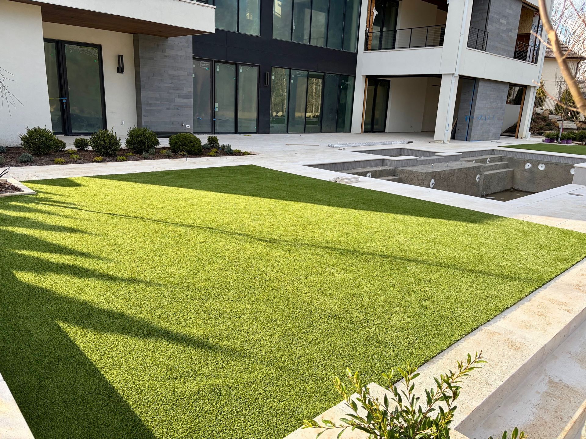 Modern house with large windows overlooking a green artificial lawn and a partially constructed stone swimming pool area.