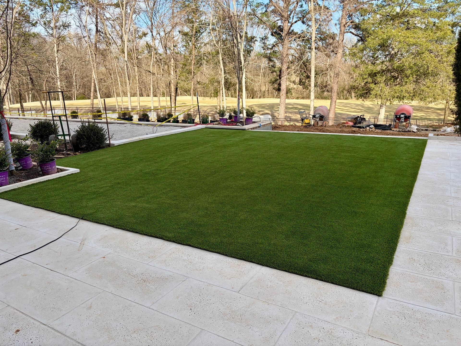 A rectangular patch of green artificial turf surrounded by light-colored stone pavers in a backyard with trees in the back.