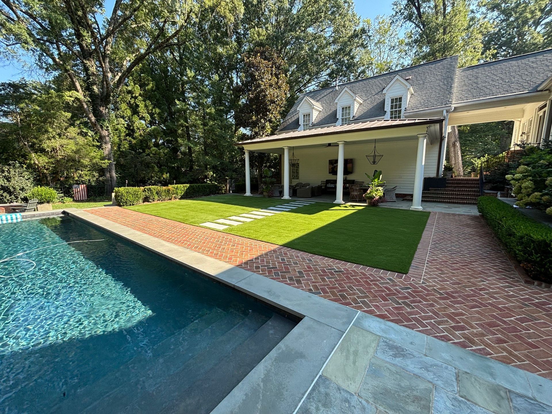 Backyard pool beside a brick patio and white house with covered porch, surrounded by trees and lawn