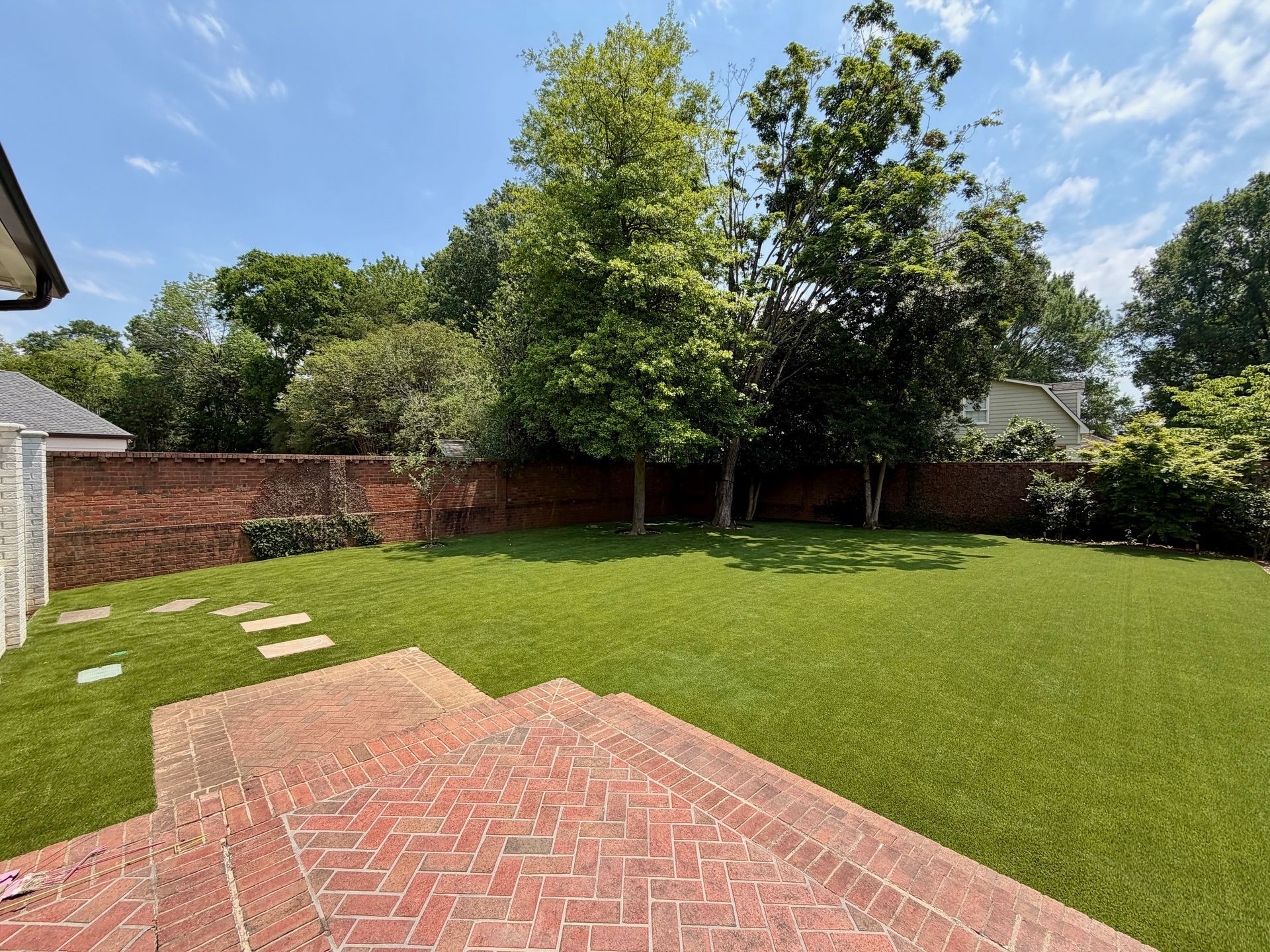 Backyard with green lawn, brick patio, and brick fence under trees and blue sky