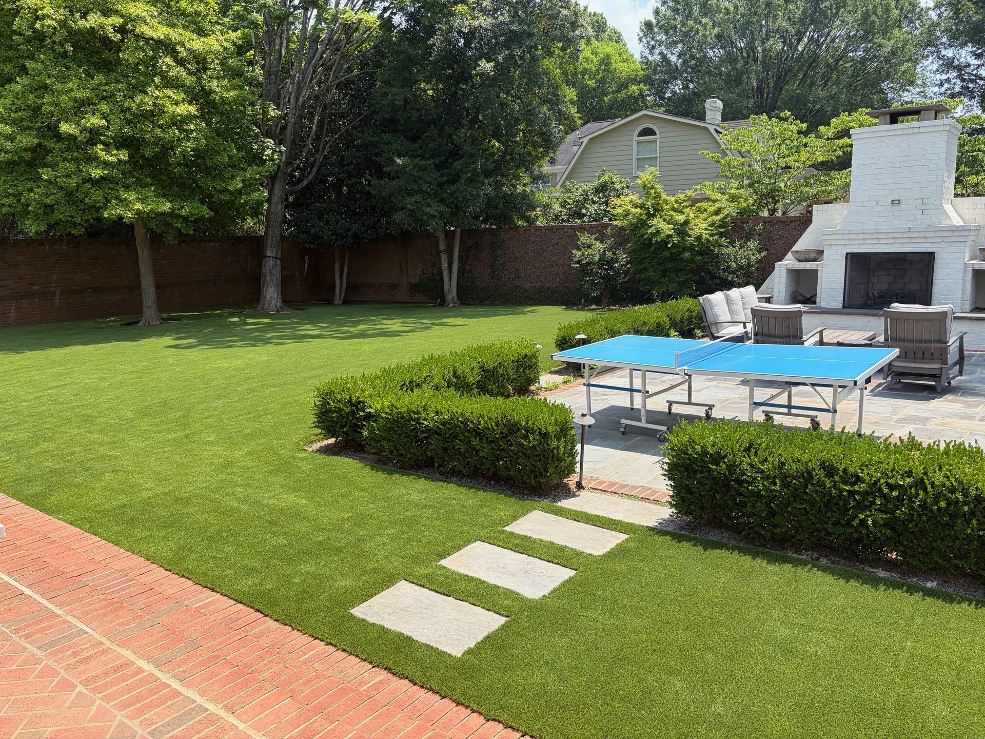 Backyard with green lawn, stone path, blue pool cover, and white outdoor fireplace beside patio seating