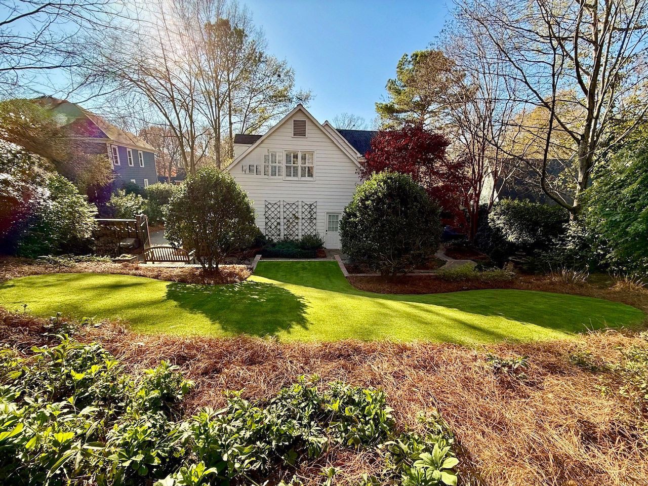 White house with a gabled roof surrounded by lush green lawn, trees, and landscaping under a sunny sky.