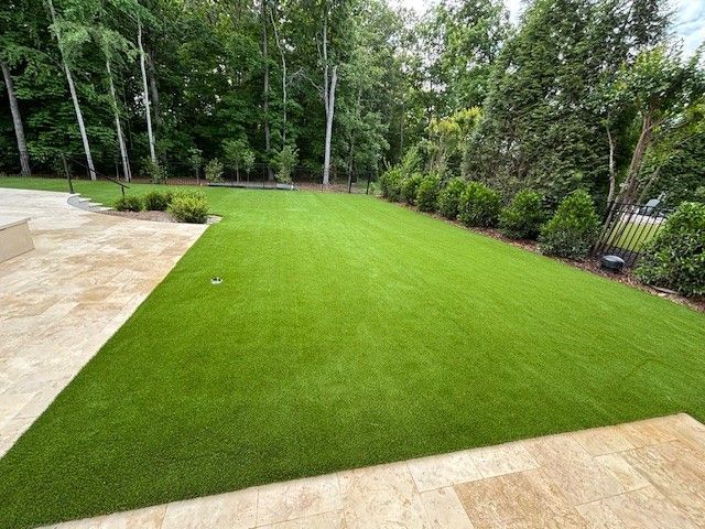 Neatly landscaped backyard with bright green lawn, stone patio, and trees along the fence line