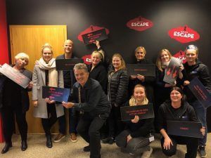 Hópur fólks situr fyrir innandyra í flóttaherbergi og heldur á dökkum skilti með rauðum „FLÓTT“ merkjum á veggnum.