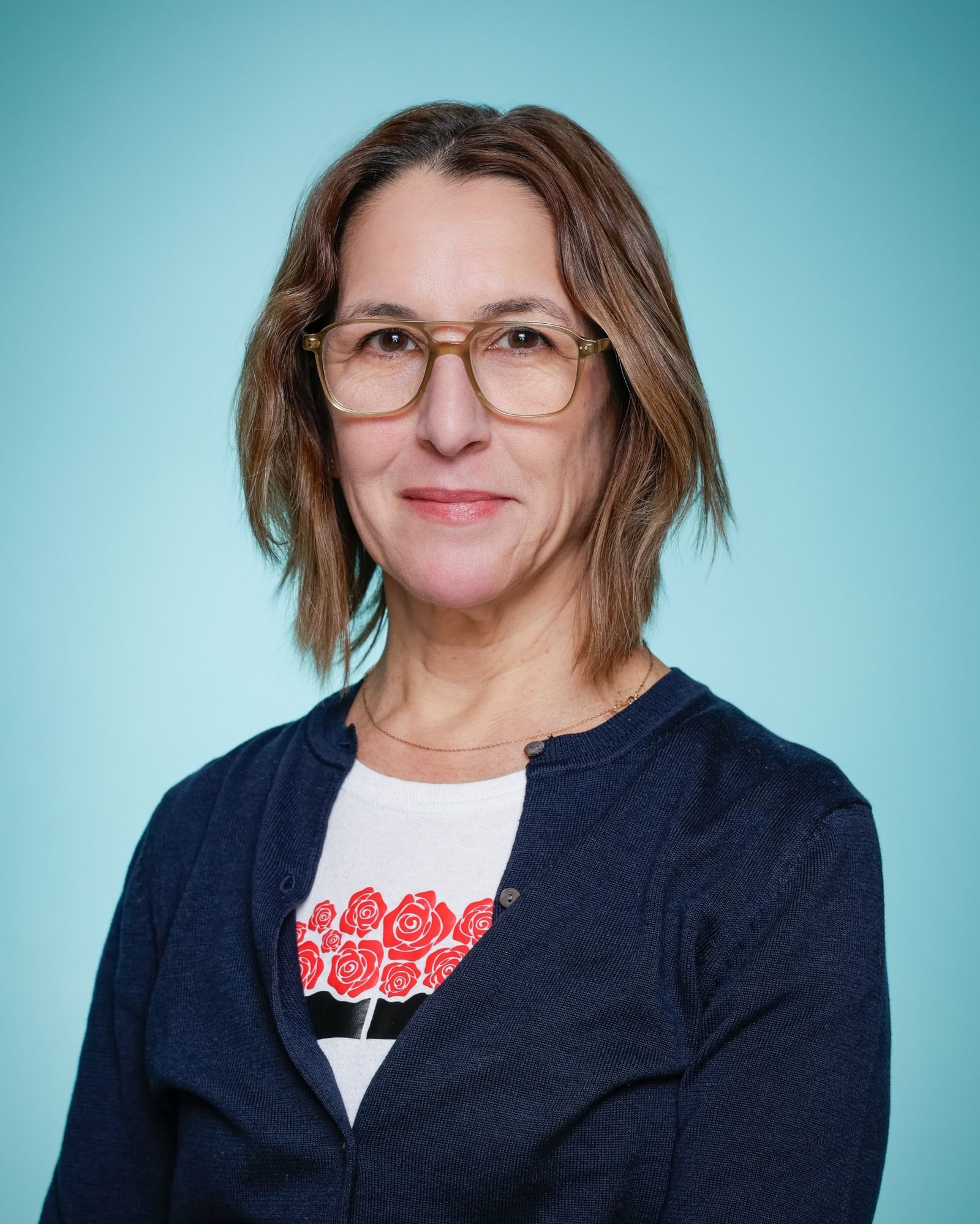 A woman wearing glasses and a necklace is smiling in front of a bookshelf.