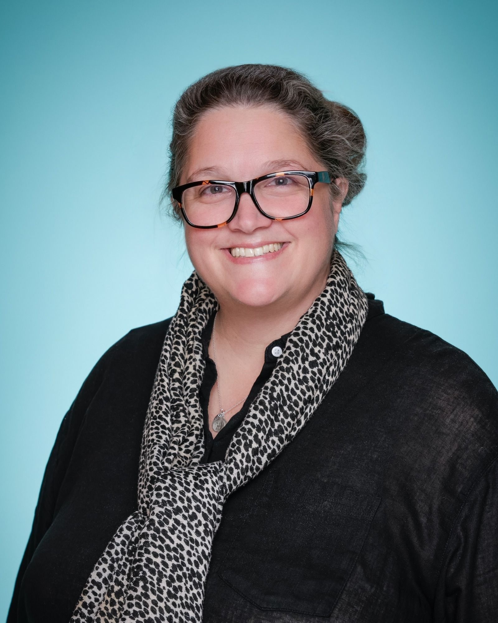 A woman wearing glasses and a black shirt is smiling in front of a bookshelf.