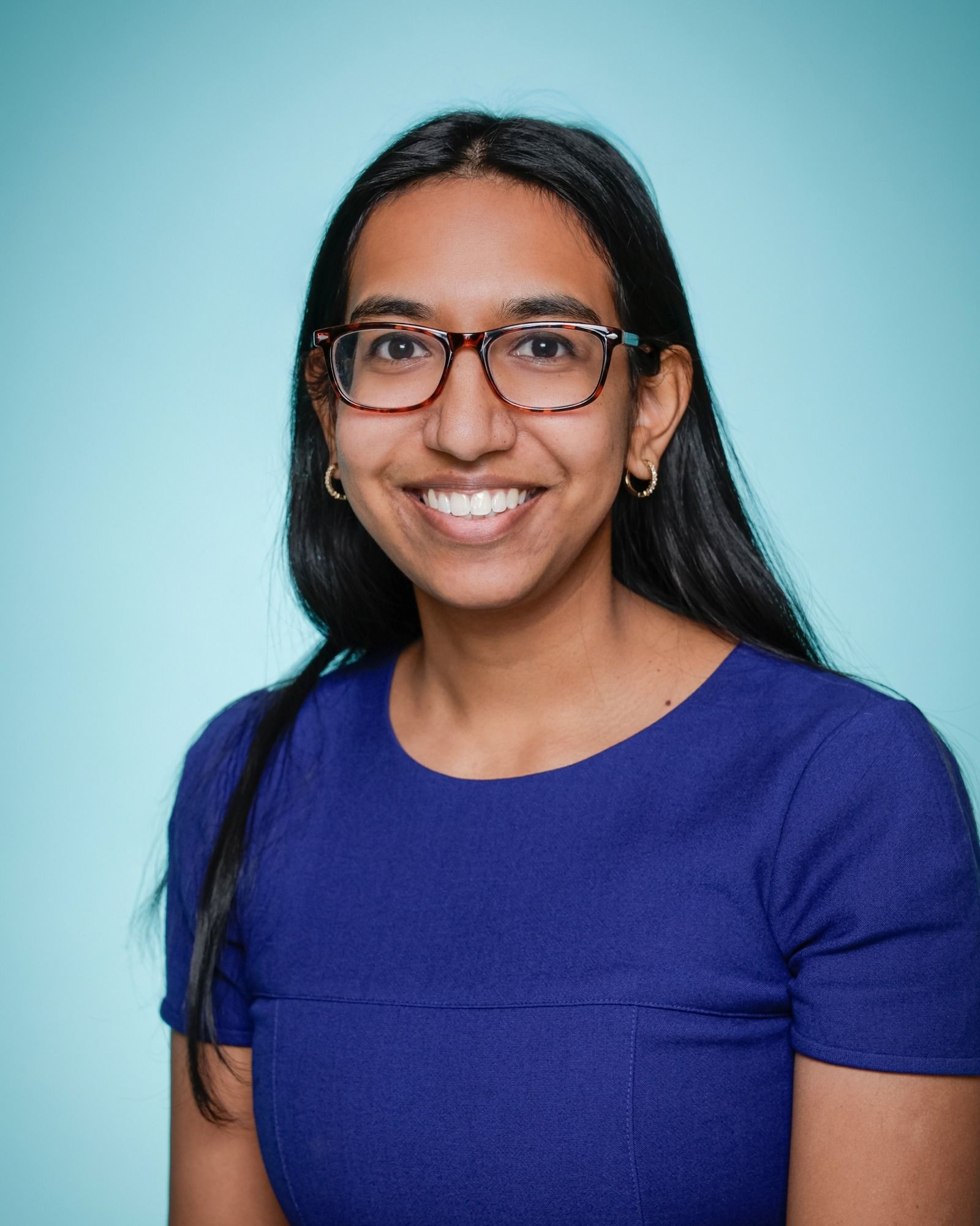 A woman wearing glasses and a plaid shirt is smiling in front of a bookshelf.