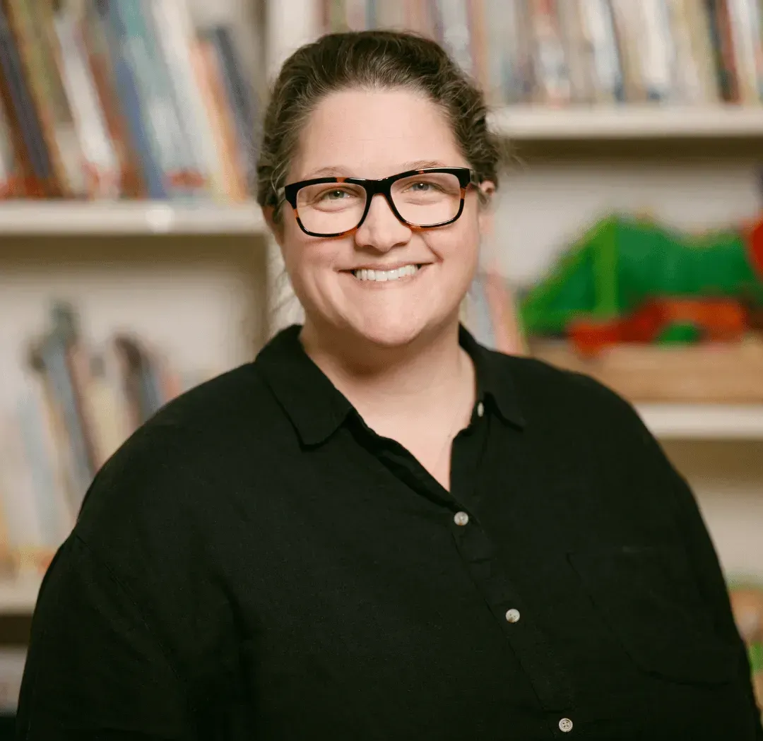 A woman wearing glasses and a black shirt is smiling in front of a bookshelf.