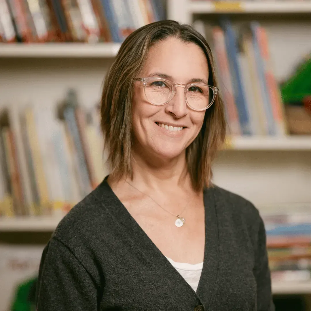 A woman wearing glasses and a necklace is smiling in front of a bookshelf.