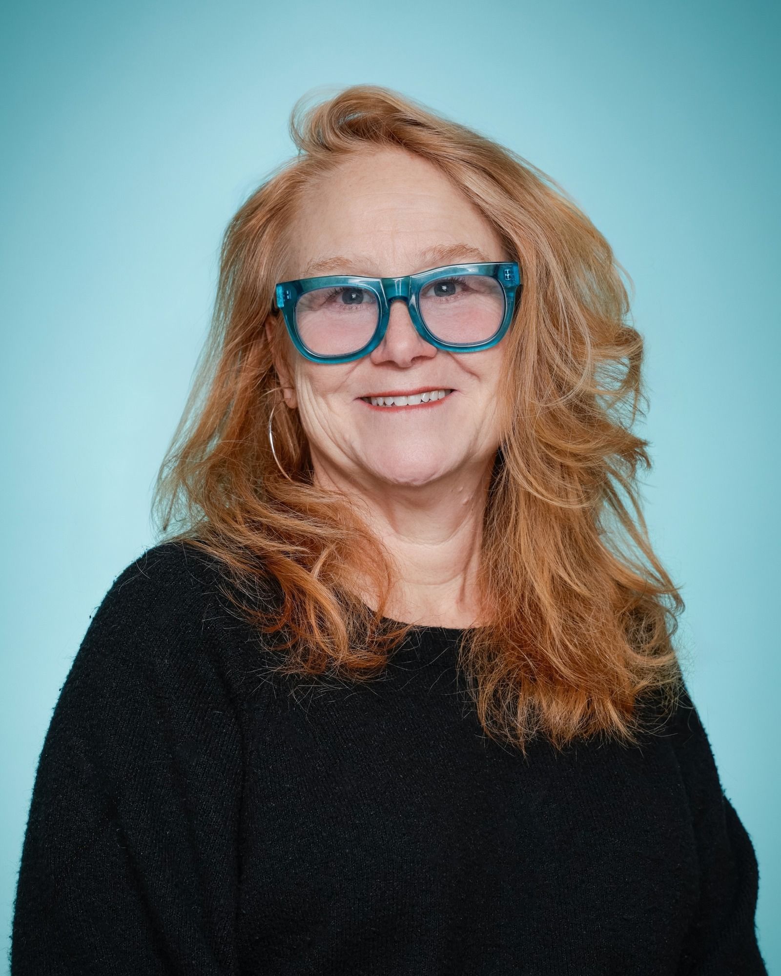 A woman wearing glasses and a blue scarf is standing in front of a bookshelf.