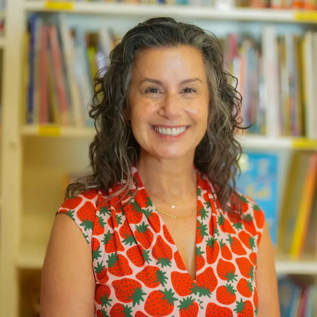 A woman in a strawberry shirt is smiling in front of a bookshelf.