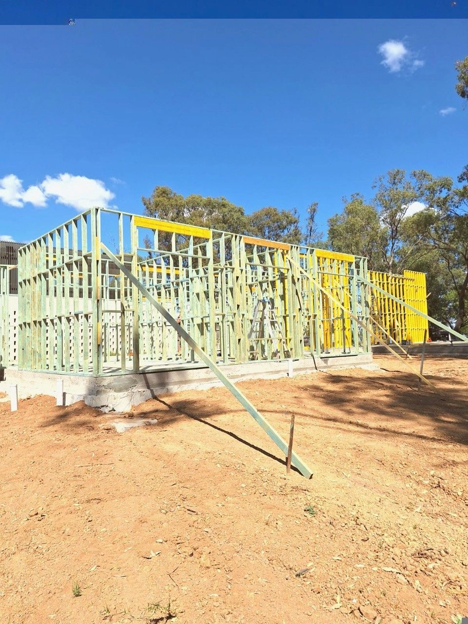 Framing of a house under construction on a dirt lot under a blue sky with trees in the background. 