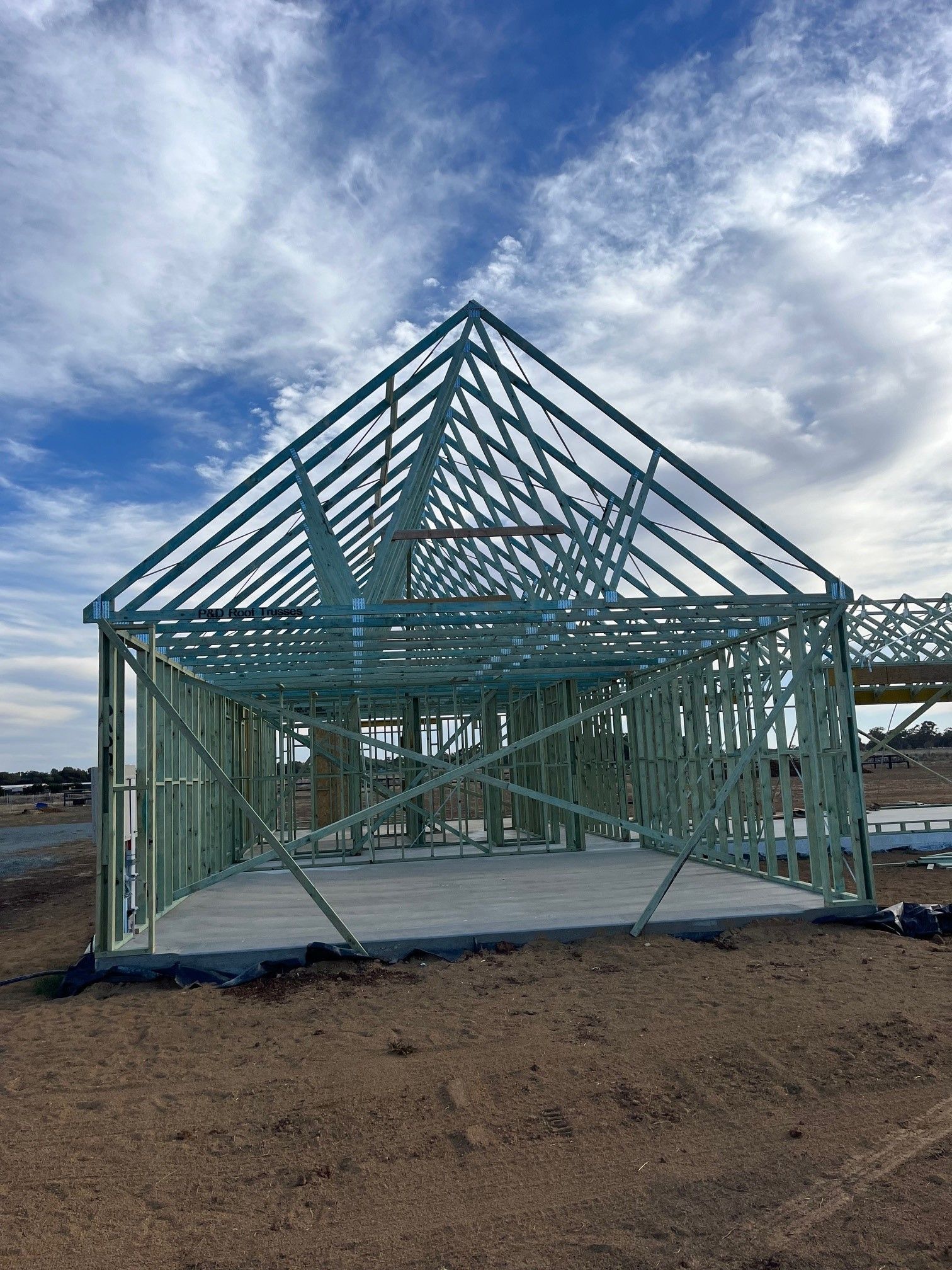 Wooden frame of a building under construction against a blue sky. The frame sits on a concrete slab in a dirt lot.