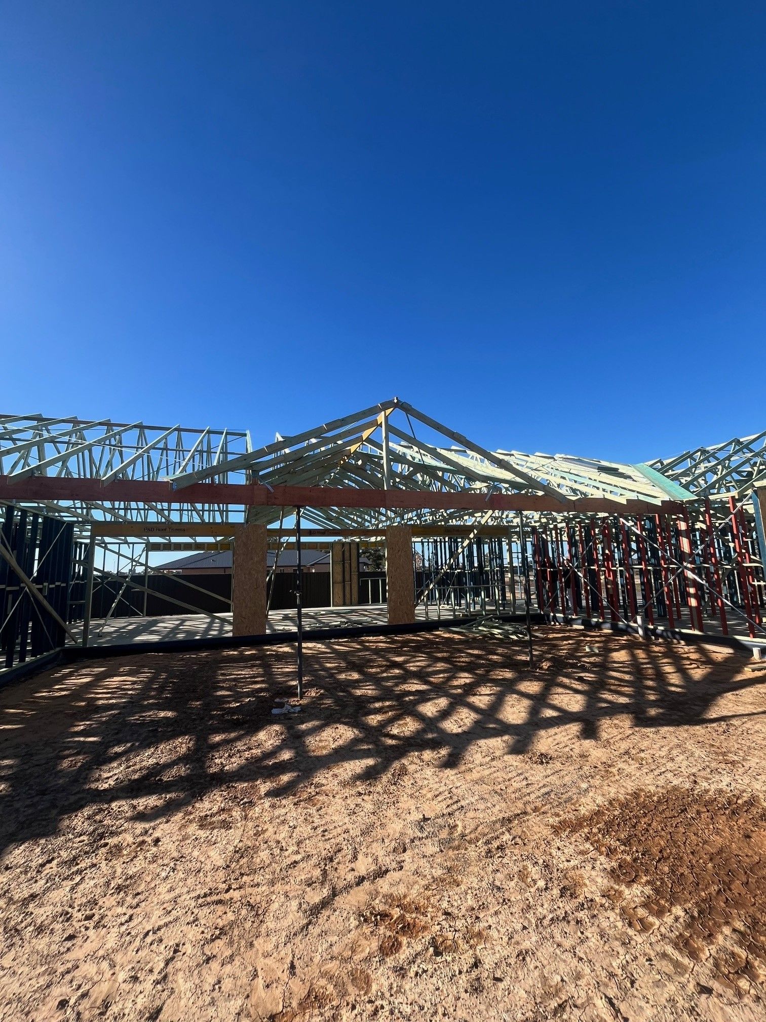 Construction site with steel frame roof and walls against a clear blue sky. Ground is dirt, with dark shadows.