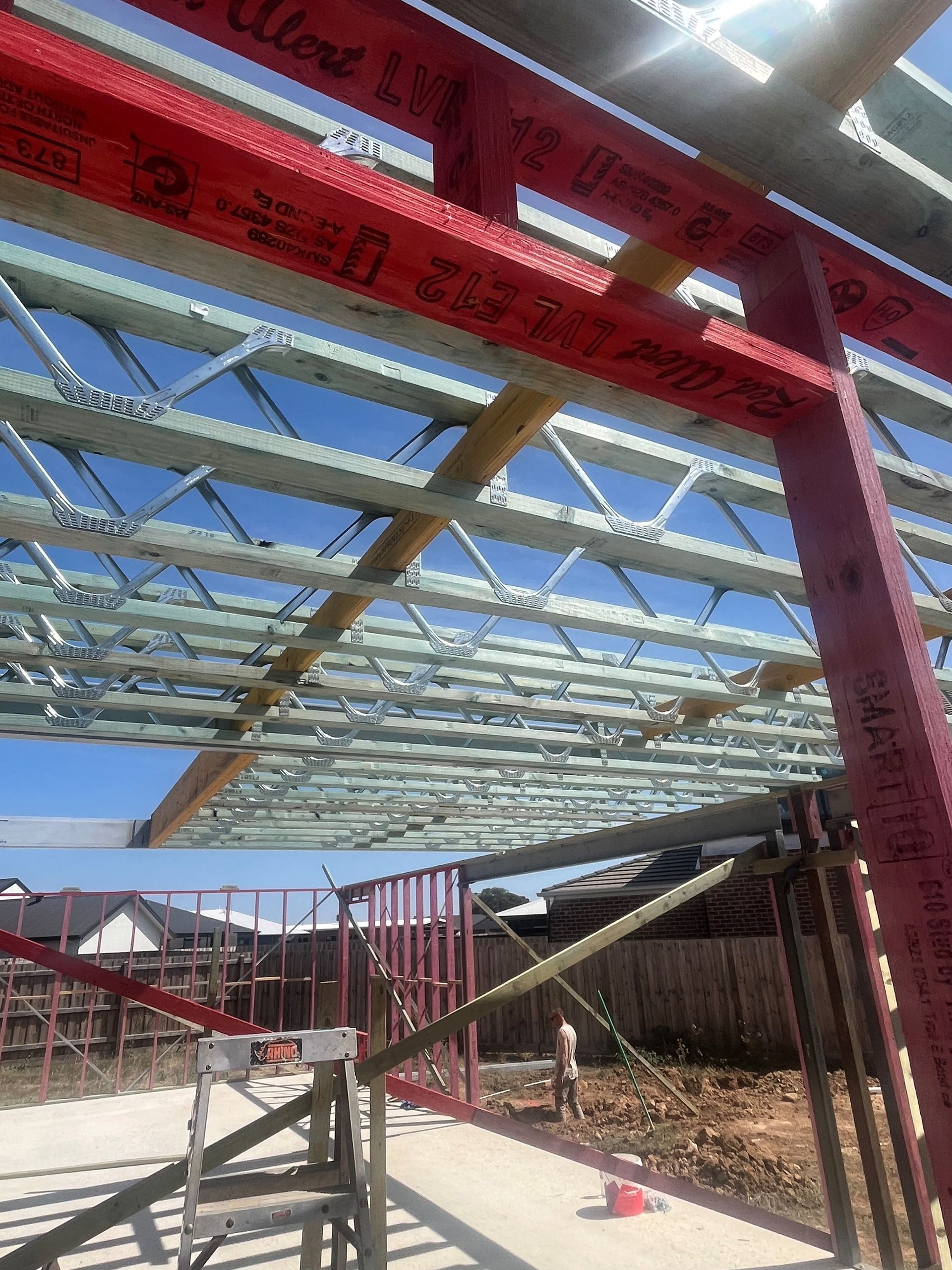 Construction site with a partially built roof structure. Red beams support metal joists against a blue sky.