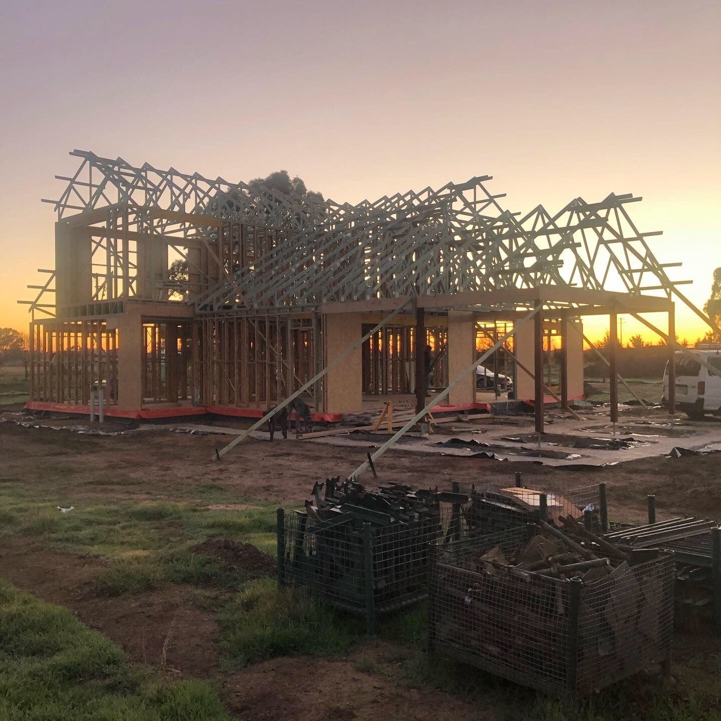 Framed house under construction at sunset, with visible wooden beams and trusses against an orange sky. 