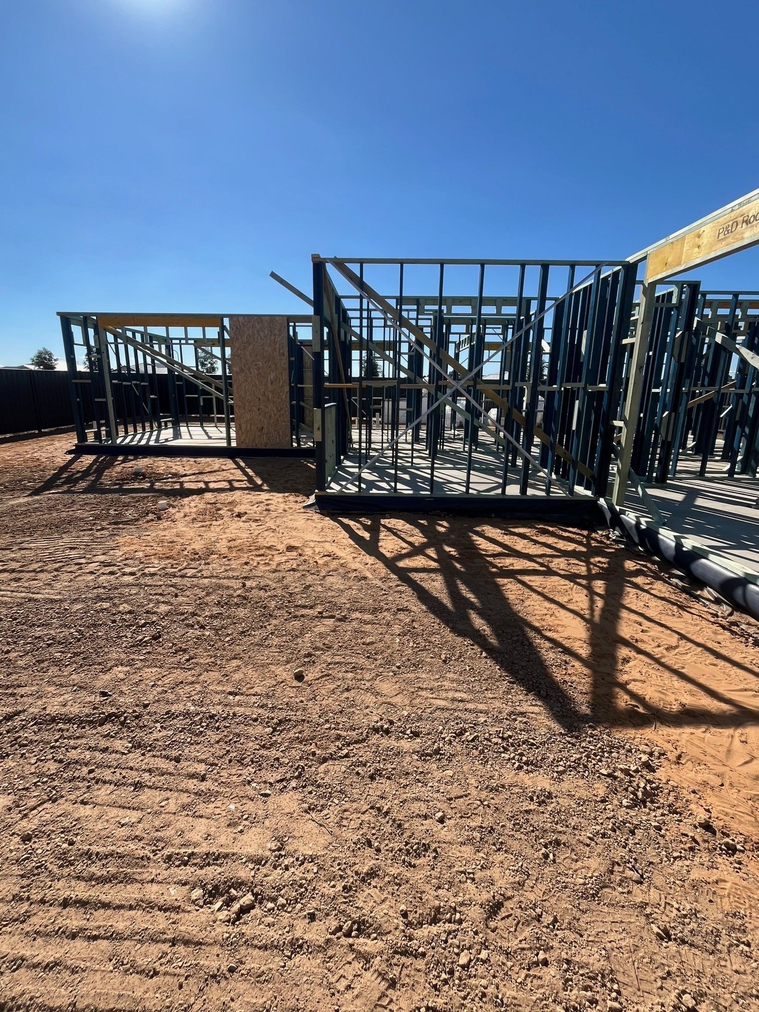 Steel frame house construction on a dirt lot under a clear, blue sky. Shadows cast by the frames stretch across the ground.