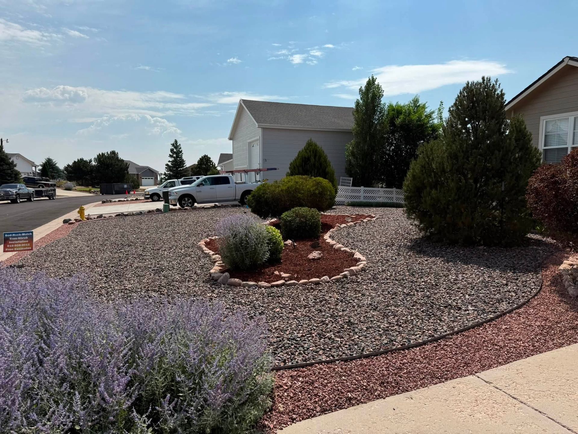 Desert-style front yard with red gravel, shrubs, and a house along a suburban street under a blue sky
