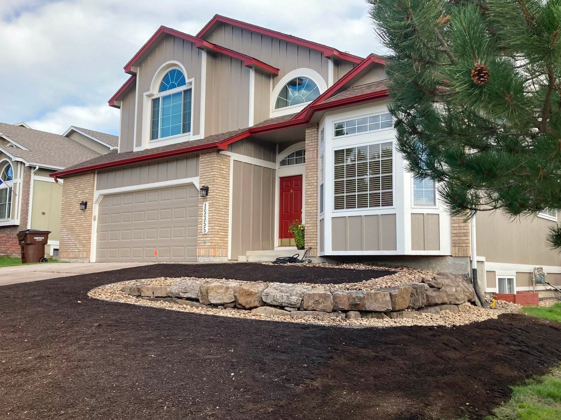 Suburban house with tan siding, red trim, a two-car garage, and a landscaped front yard.