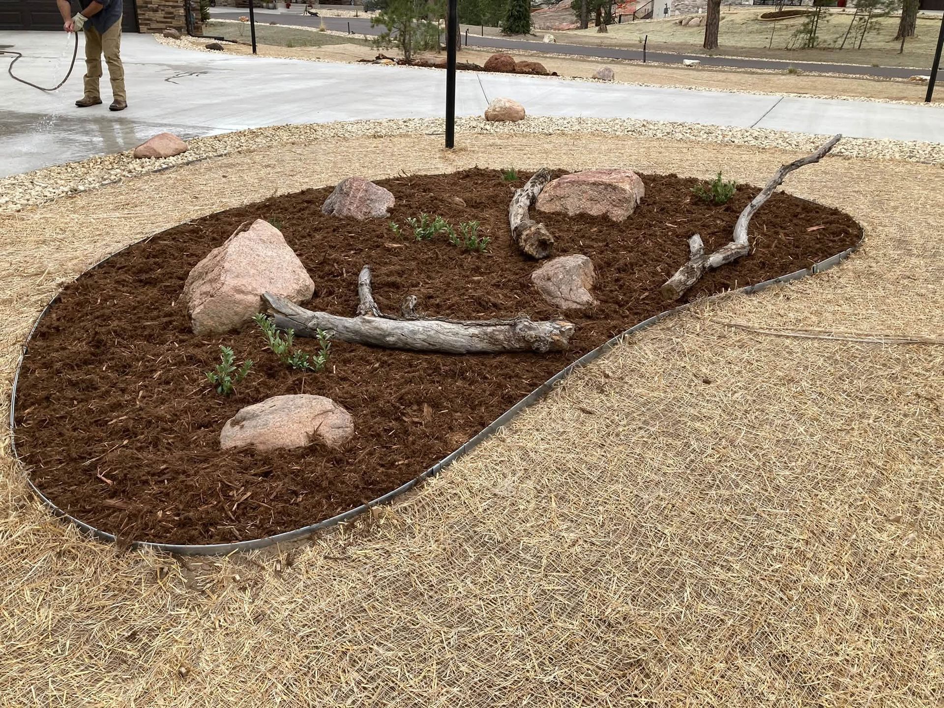Rock-lined garden bed with mulch and a curved branch on a gravel path