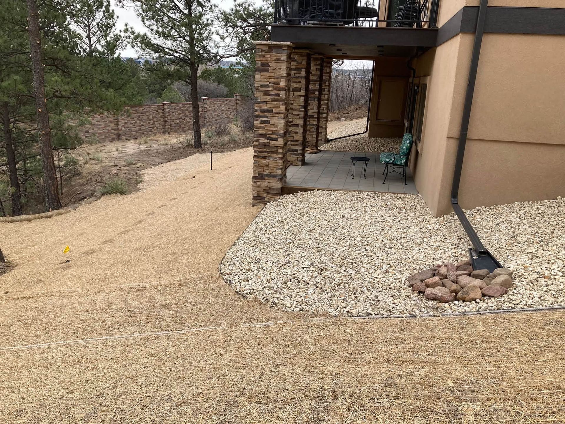 Patio beside tan house with stone pillars, gravel path, and trees in the yard