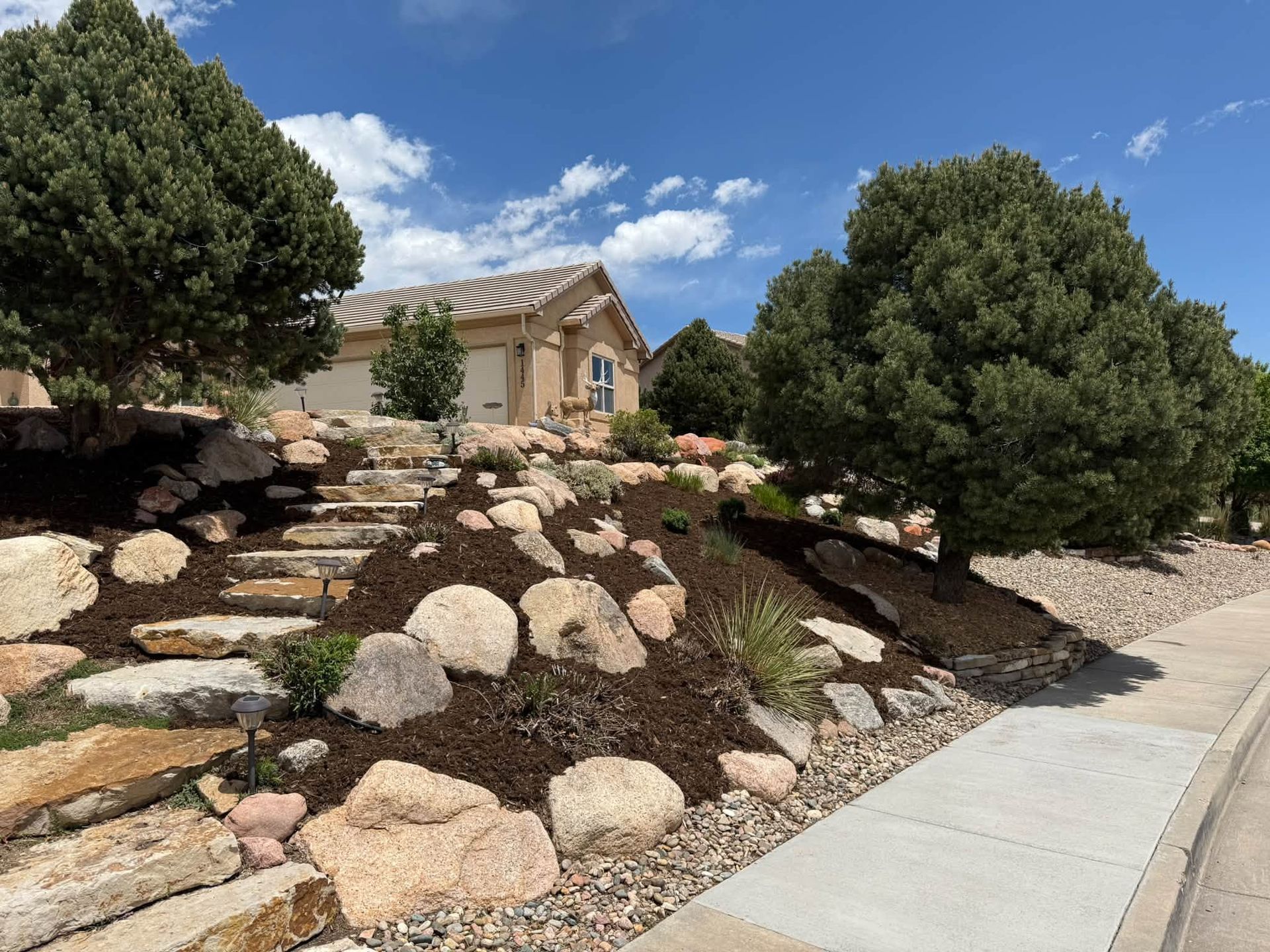 Desert landscaping with rocks and shrubs beside a sidewalk and a tan house under a blue sky.