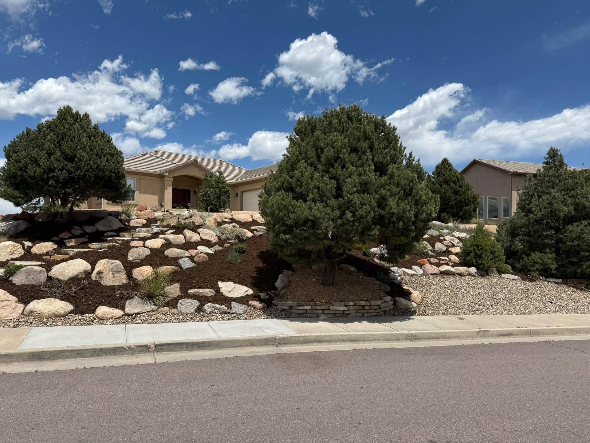 Suburban homes behind rock landscaping and trees under a blue sky with clouds