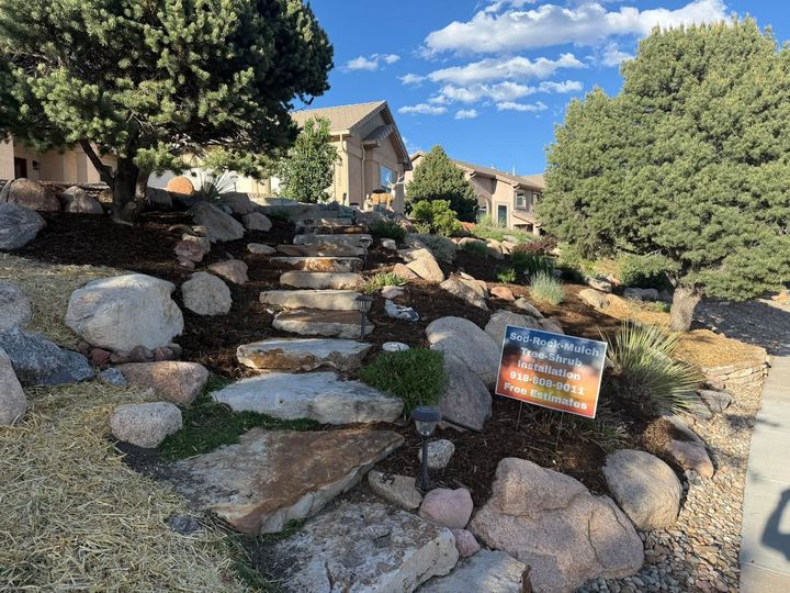 Rocky landscaped steps beside houses, with trees and a small sign under a blue sky