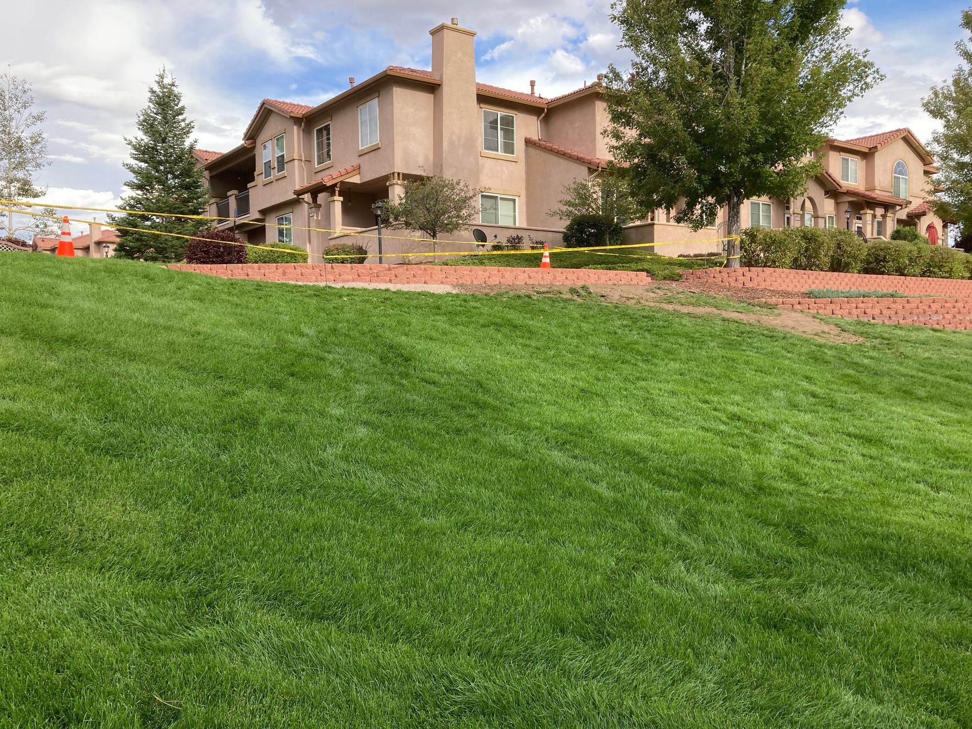Row of beige apartment buildings beside a green lawn and flower beds under a partly cloudy sky
