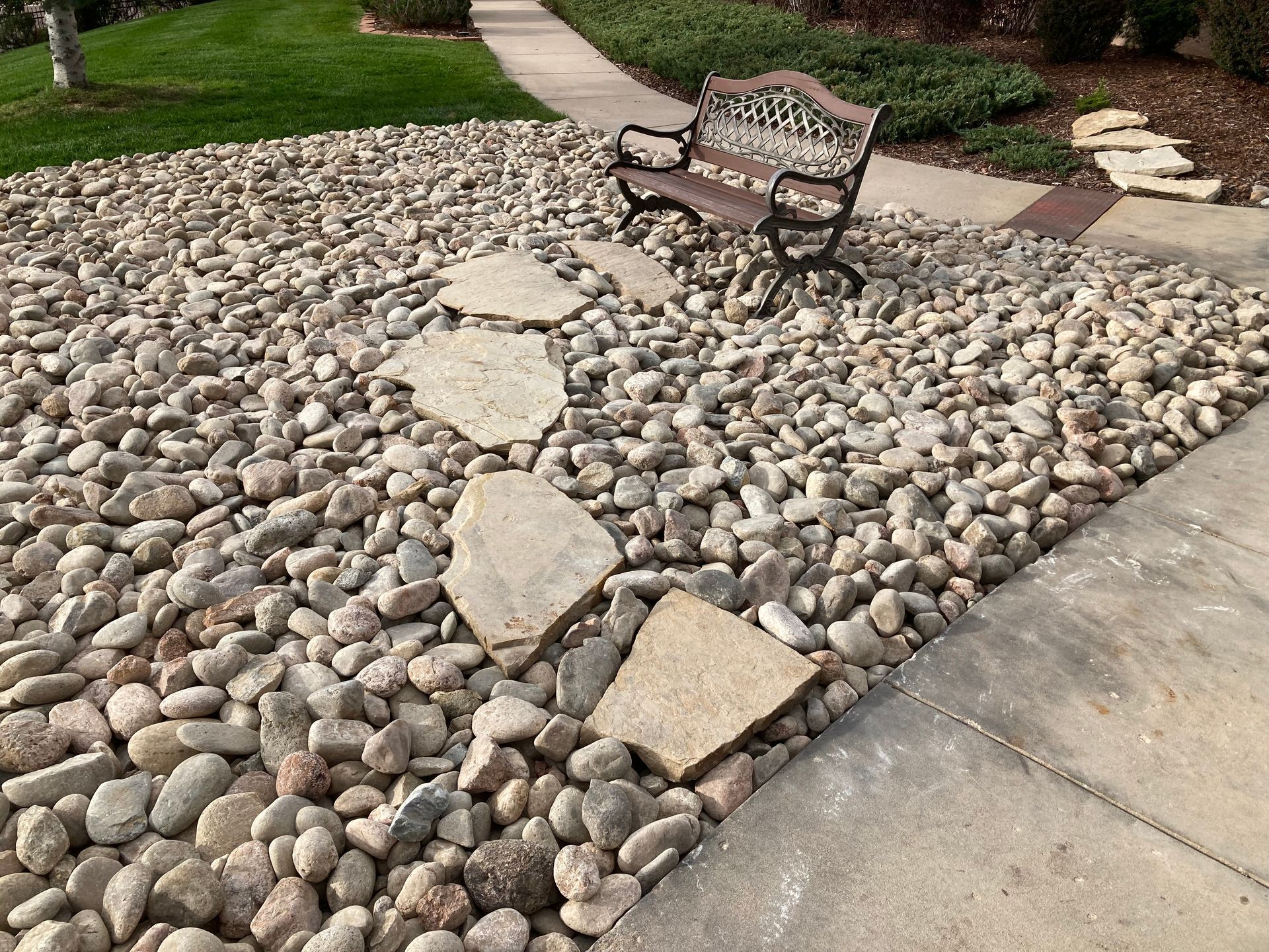 Rock-covered landscaping with stepping stones and a decorative bench beside a sidewalk