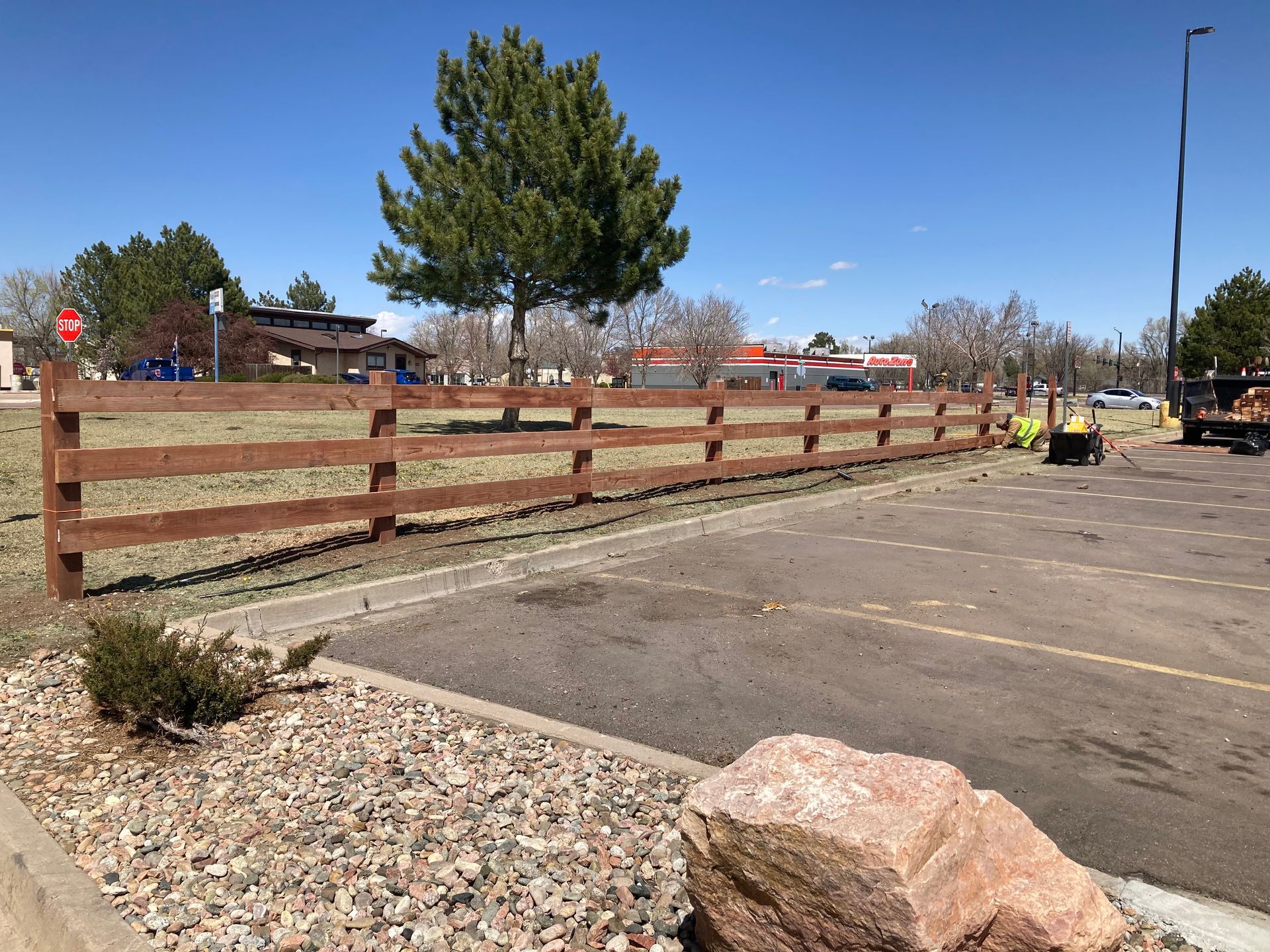 Wooden fence beside empty parking lot with trees and rocks under a clear blue sky