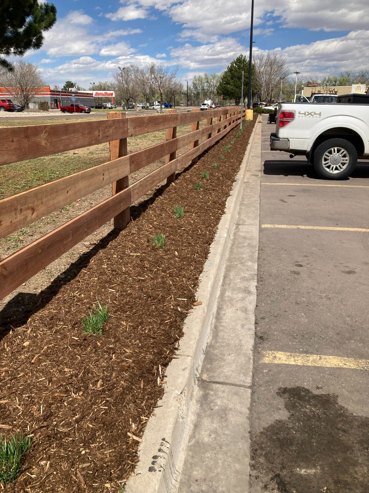 Wooden fence beside a mulched curb and parking lot with a white pickup truck under a cloudy sky.