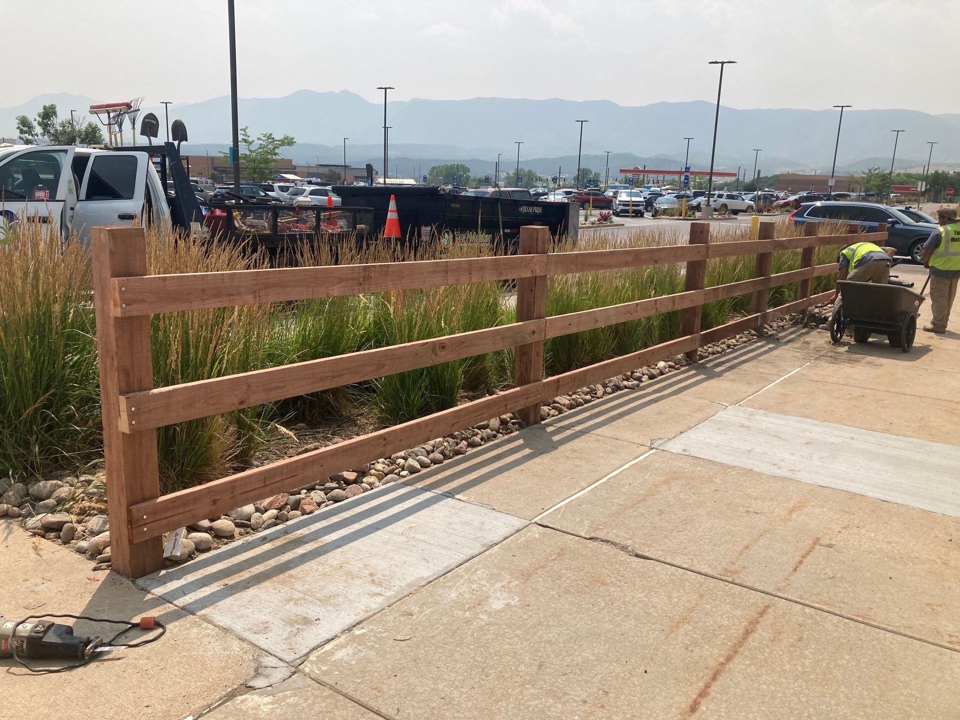 Wooden fence beside a concrete sidewalk near parked vehicles and open fields under a hazy sky