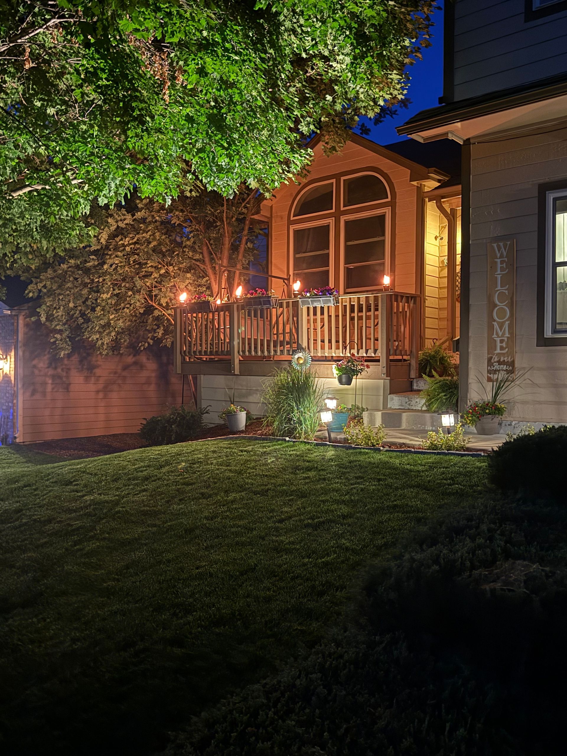 Warmly lit house porch at night with arched windows, greenery, and a lawn in front.