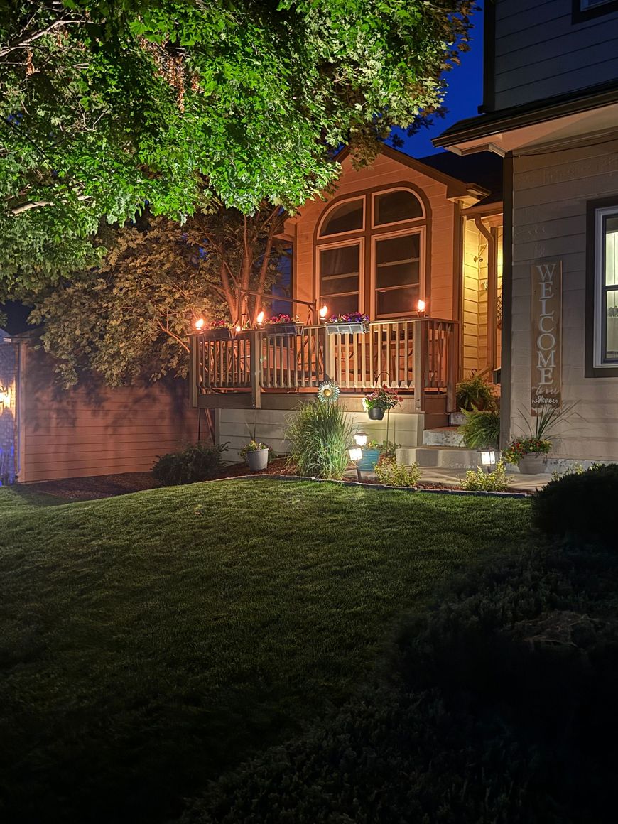 House porch lit warmly at night, with large windows, tree canopy, and a dark lawn in front.