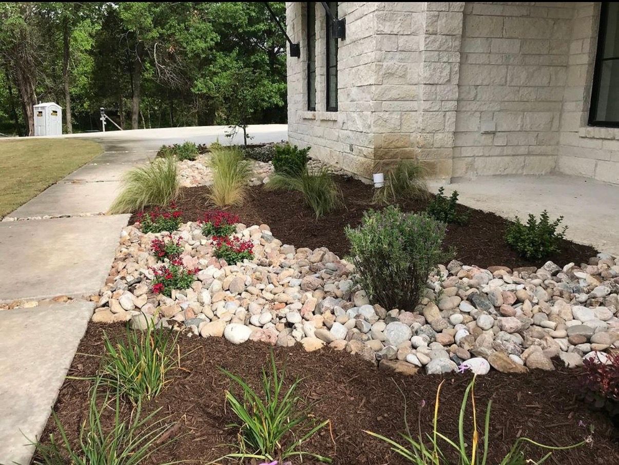 Stone house beside landscaped garden bed with flowers, shrubs, and white rocks along a walkway
