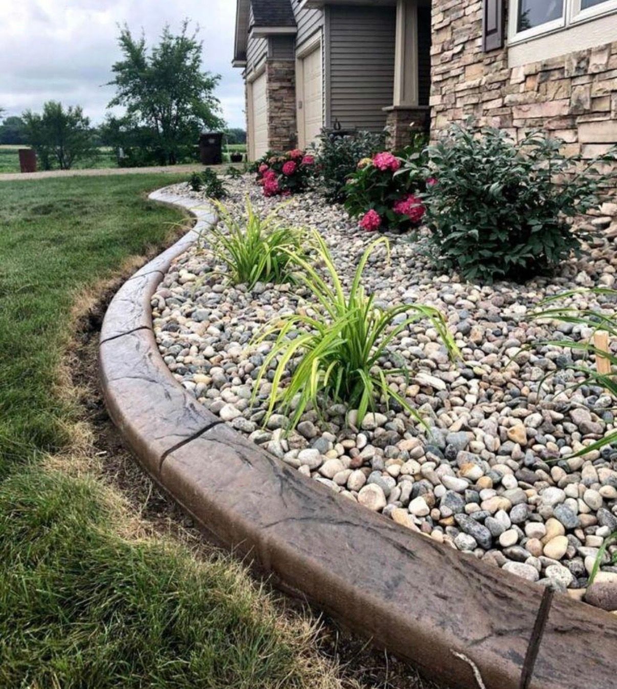 Landscaped stone border with river rocks and pink flowers beside a house and green lawn