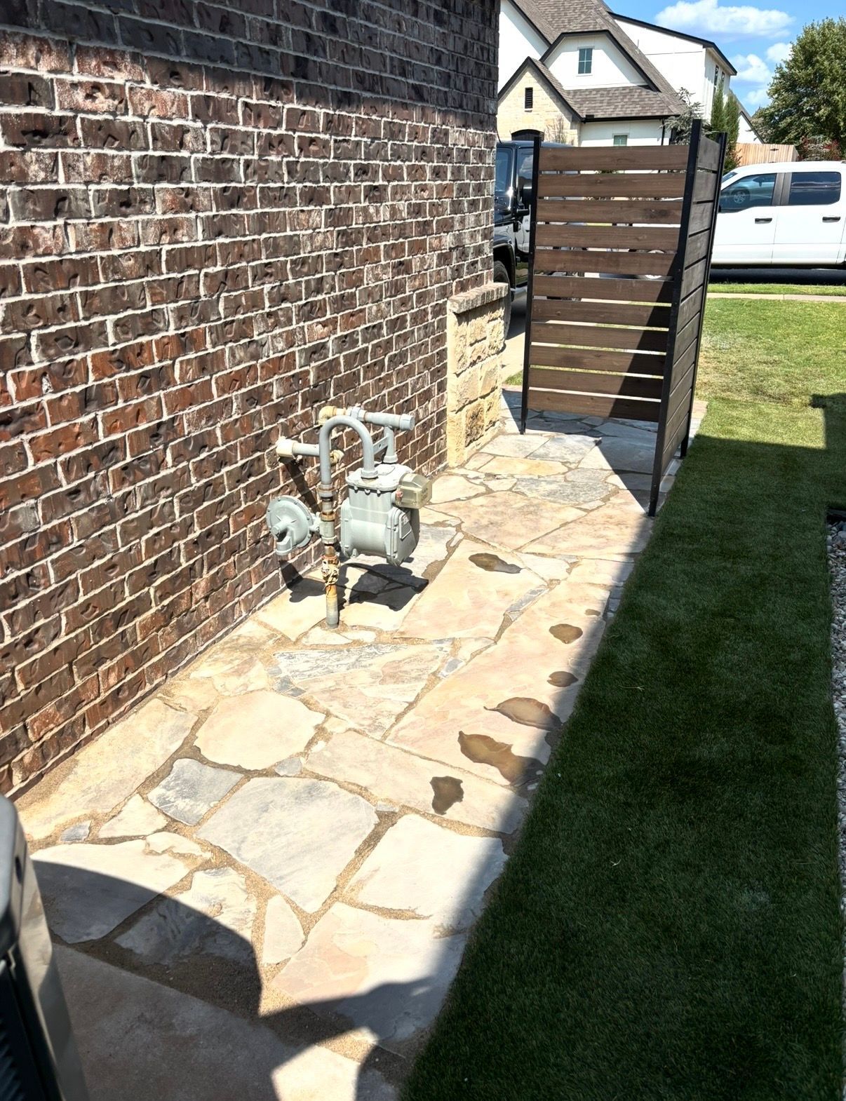 Stone walkway beside brick wall with gas meters, wooden gate, and grass lawn