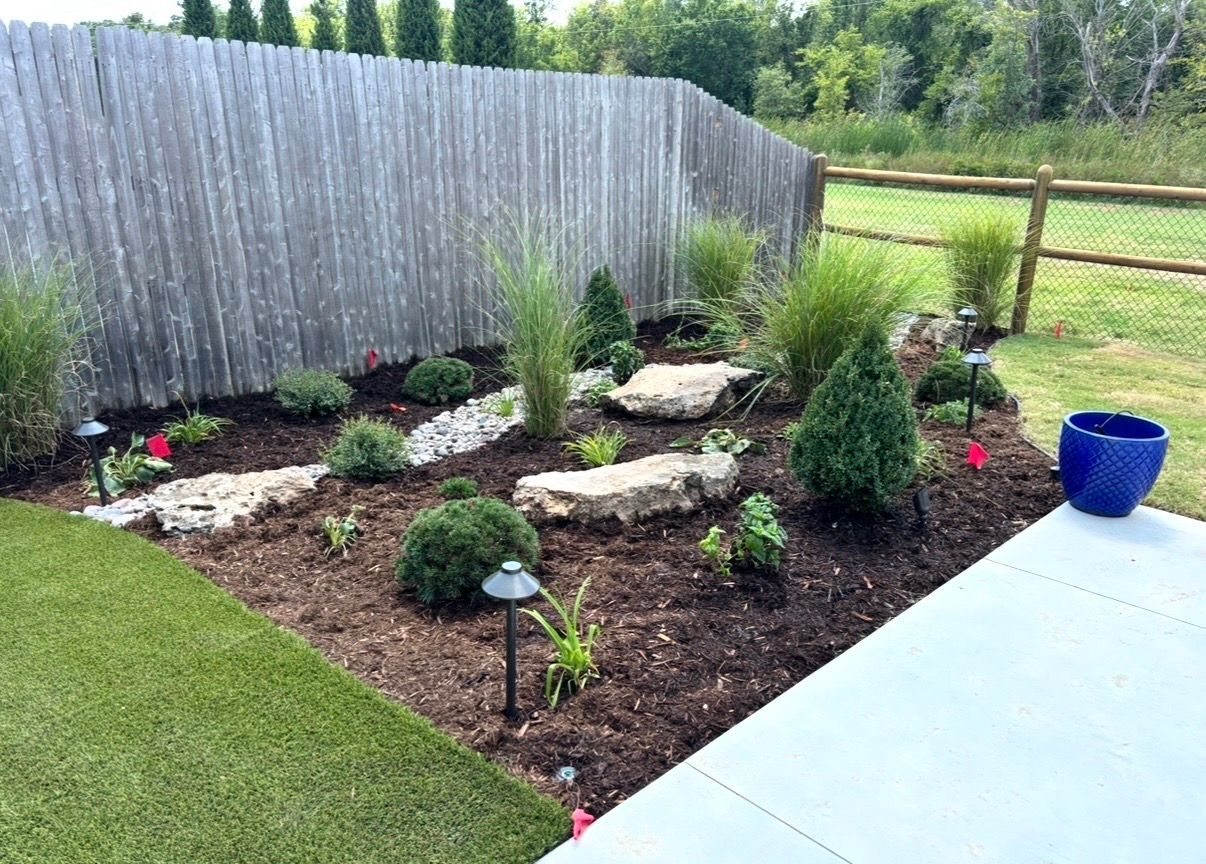 Landscaped backyard garden bed with rocks, shrubs, and a blue planter beside a patio