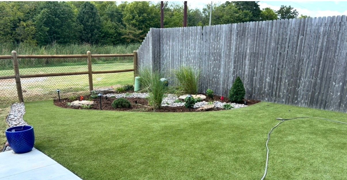 Backyard lawn with flower bed, wooden fence, and a blue planter on a patio.