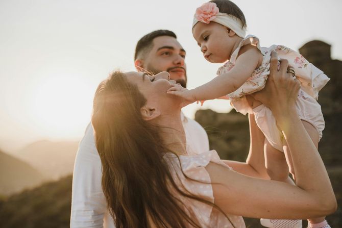 A couple holds their baby up against a sunset background, with the woman looking affectionately at the child.