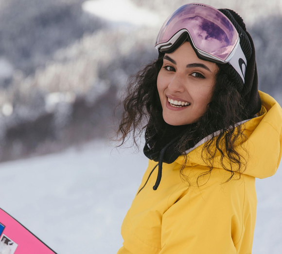 A smiling person with curly hair wears a yellow jacket and purple ski goggles against a snowy mountain background.