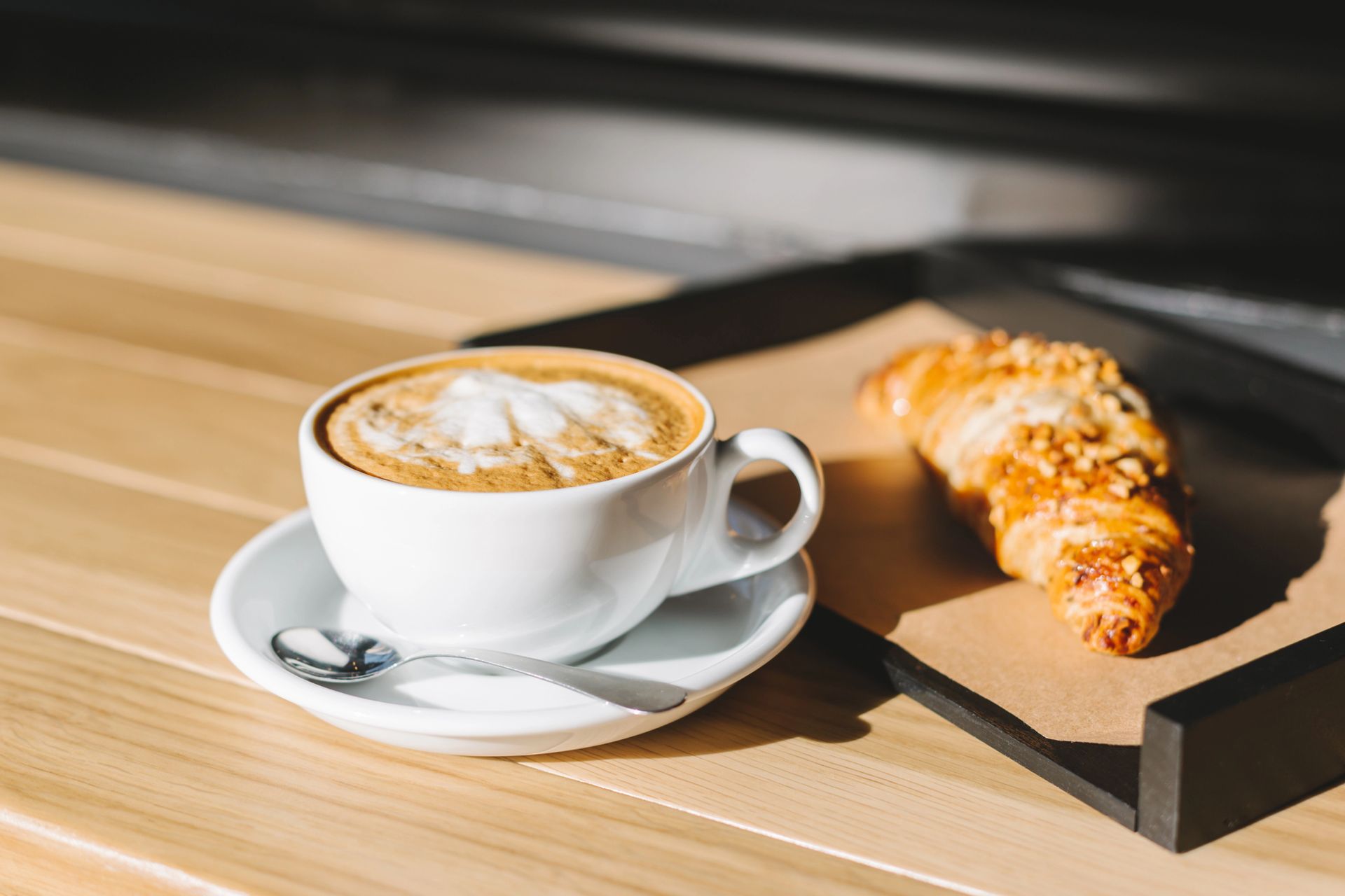 Tazza bianca da cappuccino con latte art e cornetto su un vassoio appoggiato a un tavolo di legno illuminato dal sole