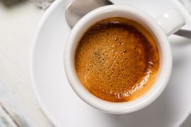 Vista dall’alto di una tazzina bianca di espresso su un piattino, con una ricca crema marrone, su un tavolo bianco.
